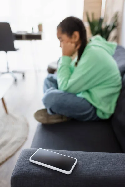 Foco seletivo do smartphone com tela em branco perto de menina afro-americana deprimida sentada no sofá, fundo borrado — Fotografia de Stock