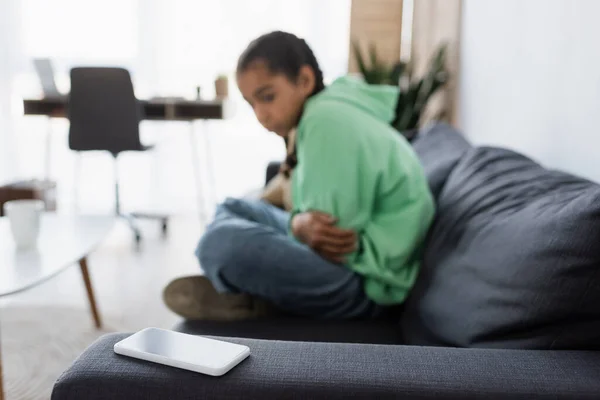Verschwommenes afrikanisch-amerikanisches Mädchen sitzt schlecht gelaunt auf der Couch und schaut auf Smartphone mit leerem Bildschirm — Stockfoto