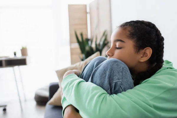 Afro-americana adolescente abrazando rodillas mientras se sienta con los ojos cerrados en casa - foto de stock