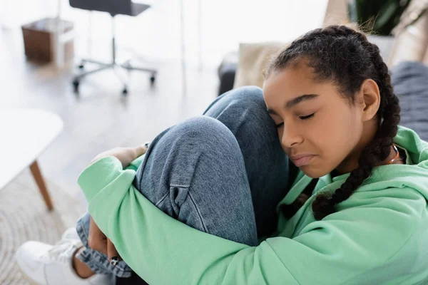 Depressed african american teen girl with closed eyes hugging knees while sitting at home — Stock Photo