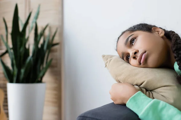 Frustrado Africano americano menina olhando para longe enquanto deitado no travesseiro perto de planta turva — Fotografia de Stock