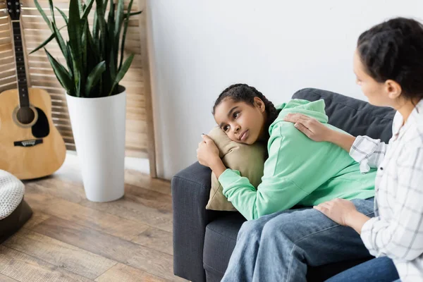 Borrosa africana americana madre calmando molesto hija abrazando almohada y mirando hacia otro lado - foto de stock