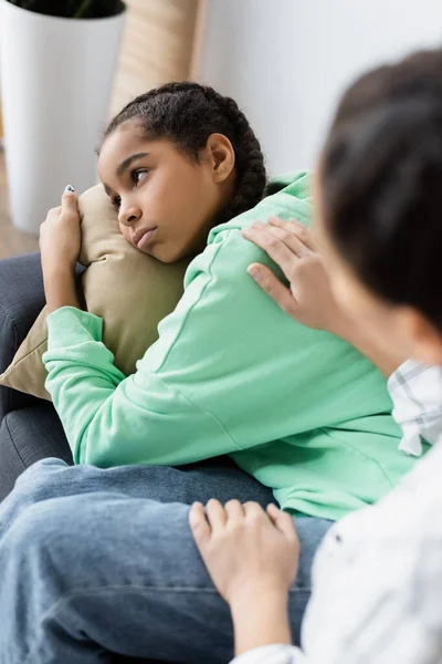 Verschwommene afrikanisch-amerikanische Frau beruhigt depressive Tochter, die auf Kissen liegt und wegschaut — Stockfoto