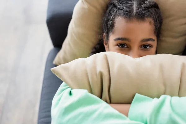 Draufsicht auf deprimierte Afroamerikanerin, die ihr Gesicht mit Kissen verdeckt, während sie zu Hause liegt — Stockfoto