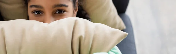 Vista superior de la chica afroamericana de mal humor mirando a la cámara mientras cubre la cara con almohada, pancarta - foto de stock