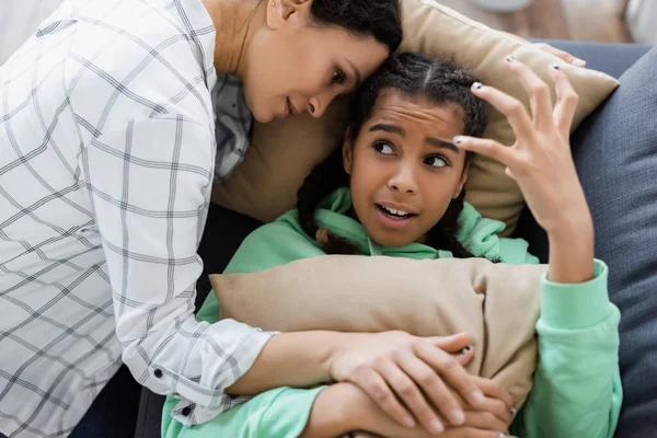 African american woman calming irritated daughter gesturing while lying on couch with pillows — Stock Photo