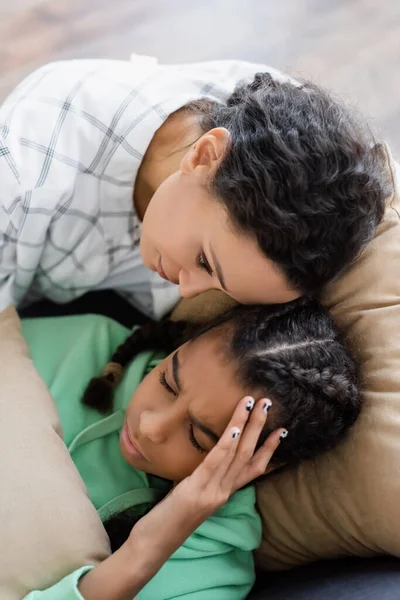 Vue de dessus de la femme afro-américaine apaisant adolescente couchée avec les yeux fermés et souffrant de maux de tête — Photo de stock