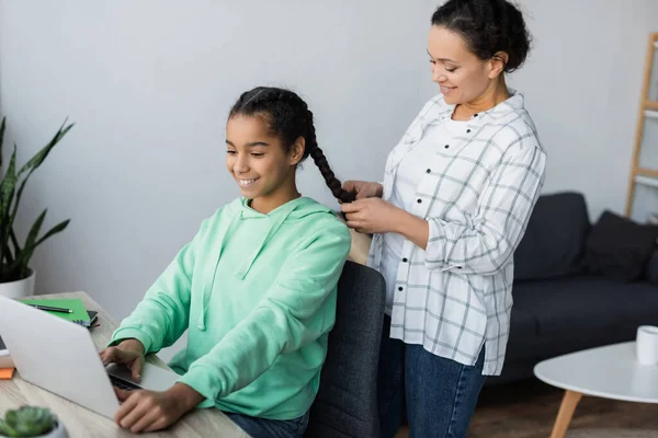 Smiling african american woman braiding hair of teenage daughter using laptop while studying at home — Stock Photo