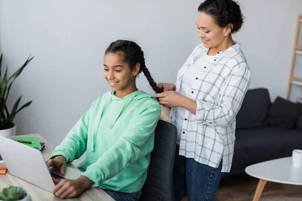 Sonriente mujer afroamericana trenzando el pelo de hija adolescente usando el ordenador portátil mientras estudia en casa - foto de stock