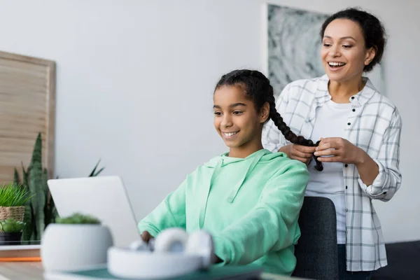 Fröhliche afrikanisch-amerikanische Frau Flechten Haare der Tochter Hausaufgaben auf Laptop — Stockfoto