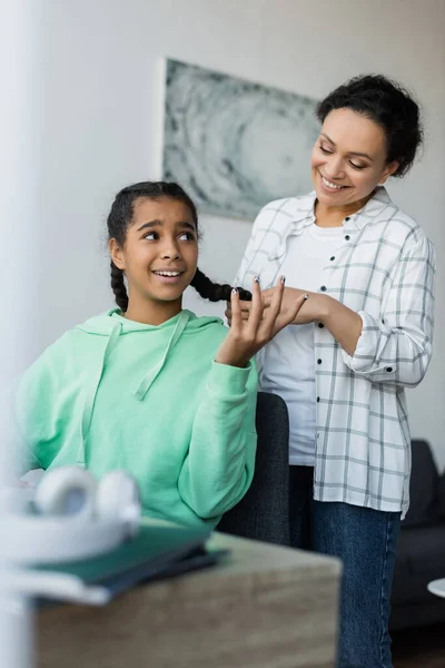 Cheerful african american woman braiding hair of discouraged daughter gesturing while sitting at desk — Stock Photo