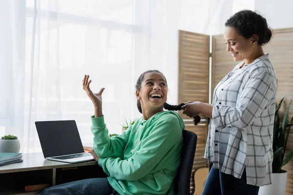 Aufgeregtes afrikanisch-amerikanisches Mädchen gestikuliert in der Nähe von Laptop mit leerem Bildschirm und Mutter, die ihre Haare geflochten hat — Stockfoto