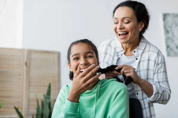 Cheerful african american woman braiding hair of laughing daughter covering mouth with hand — Stock Photo