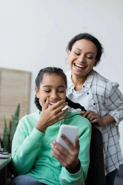 Mulher americana africana alegre trançando o cabelo da filha rindo olhando para o telefone móvel — Fotografia de Stock