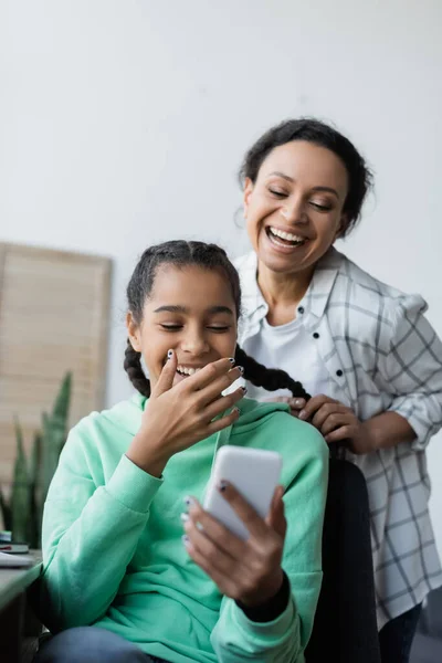 Gai afro-américaine femme tressage cheveux de rire fille regardant téléphone mobile — Photo de stock