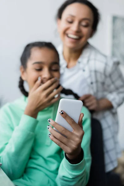 Happy african american woman braiding hair of teenage daughter laughing while looking at smartphone — Stock Photo