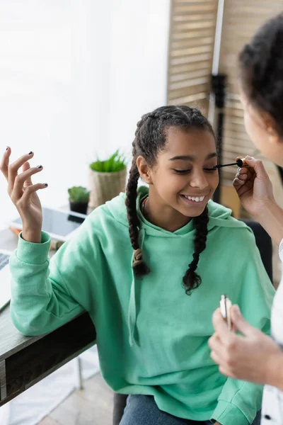 Blurred african american woman applying mascara on eyelashes of happy teen daughter — Stock Photo