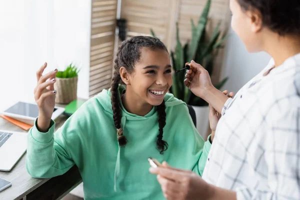 Borrosa africana americana mujer sosteniendo rímel mientras haciendo rímel a alegre hija - foto de stock