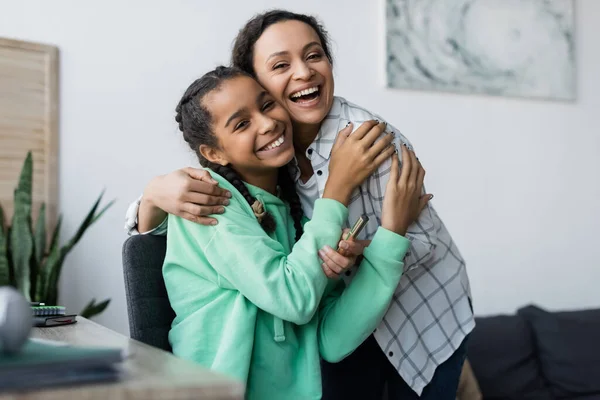 Glücklich afrikanisch-amerikanische Frau hält Wimperntusche, während sie Teenager-Tochter zu Hause umarmt — Stockfoto