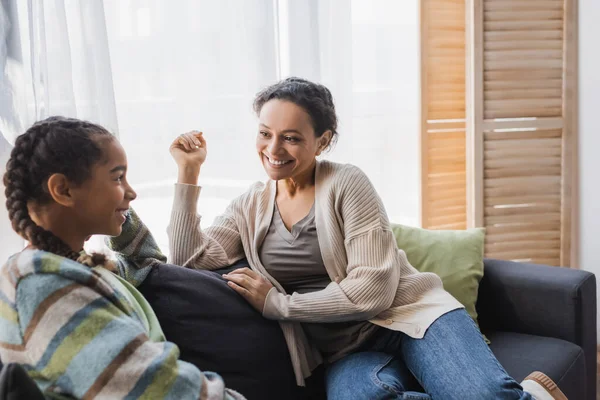 Smiling african american woman talking with teenage daughter on couch at home — Stock Photo