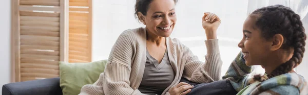 Pleased african american mother and teenage daughter talking on couch at home, banner — Stock Photo