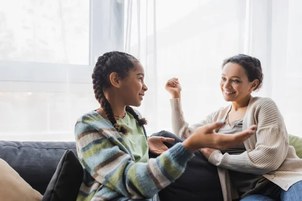Teenage african american girl pointing with hand while talking to smiling mother on sofa — Stock Photo