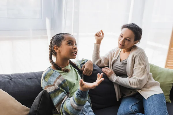 Africano americano adolescente apuntando con la mano mientras hablando con mamá en sofá en casa - foto de stock