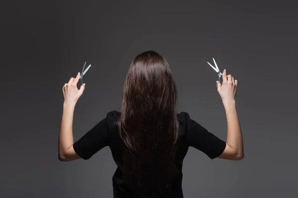 Rückansicht einer jungen Frau mit langen, glänzenden Haaren, die eine Schere isoliert auf dunkelgrau hält — Stockfoto