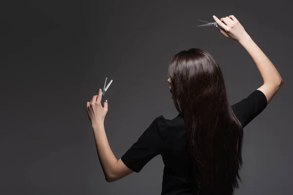 Visão traseira da mulher com cabelo longo brilhante segurando tesoura isolada no cinza escuro — Fotografia de Stock