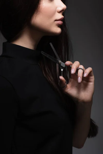 Vista recortada de mujer en cuello alto negro sosteniendo tijeras cerca de pelo largo aislado en gris oscuro - foto de stock
