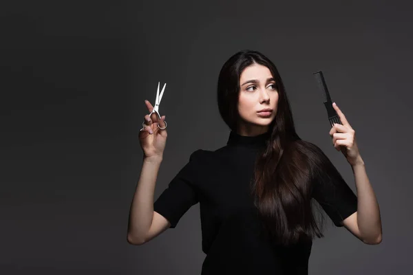 Молода жінка з довгим блискучим волоссям тримає ножиці і гребінці ізольовані на темно-сірому — стокове фото