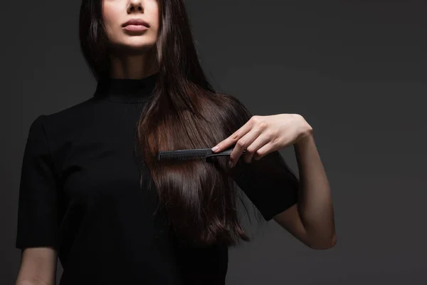 Обрезанный вид молодой женщины, расчесывающей длинные блестящие волосы расческой, изолированной на темно-серой — стоковое фото