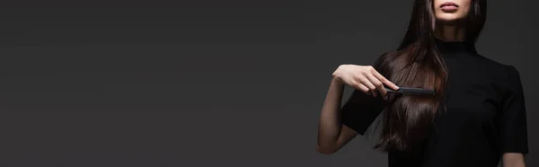 Обрезанный вид молодой женщины, расчесывающей длинные блестящие волосы расческой, изолированной на темно-сером, баннер — стоковое фото
