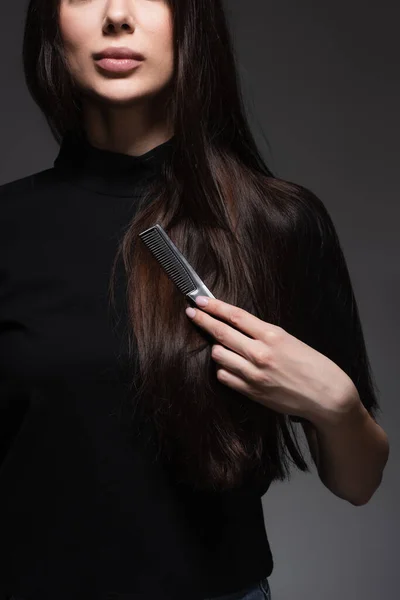 Vue partielle de la jeune femme brossant de longs cheveux brillants avec peigne isolé sur gris foncé — Photo de stock