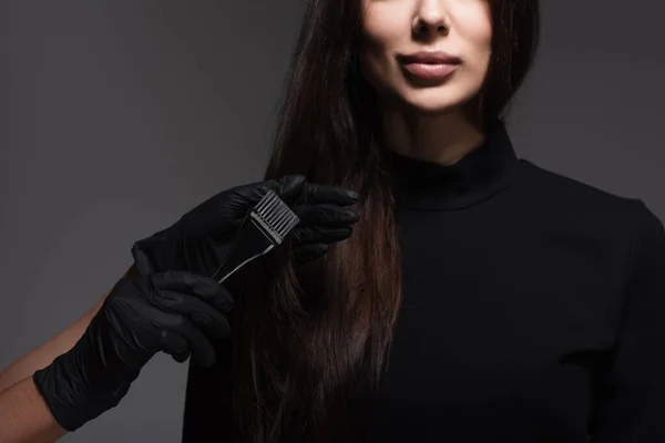 Vista ritagliata del parrucchiere tenendo pennello vicino alla donna con i capelli castani isolati sul grigio scuro — Foto stock