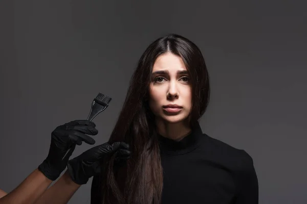 Hairstylist holding brush near sad woman with brown hair isolated on dark grey — Stock Photo