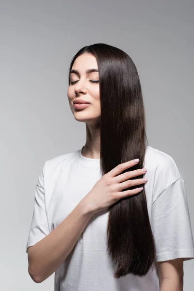 Mujer complacida tocando el pelo largo liso aislado en gris - foto de stock