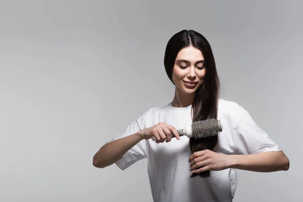 Femme heureuse brossage cheveux longs lisses avec brosse thermique isolé sur gris — Photo de stock