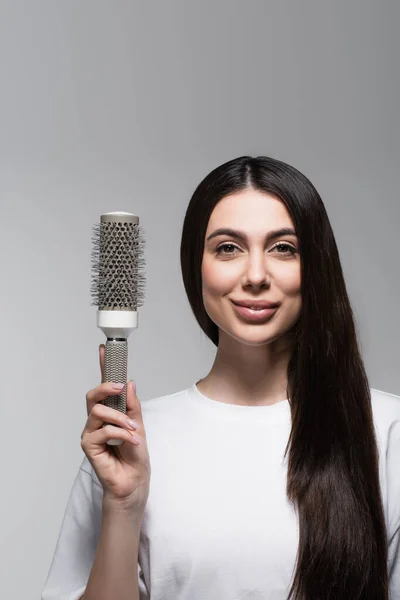 Mujer complacida con cabello largo liso sosteniendo cepillo térmico aislado en gris - foto de stock