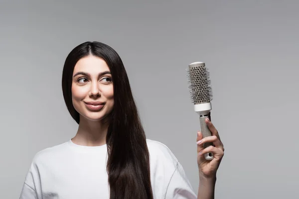 Femme heureuse avec les cheveux longs lisses regardant brosse thermique isolé sur gris — Photo de stock