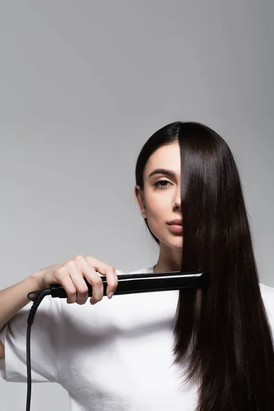 Jeune femme brune utilisant lisseur de cheveux isolé sur gris — Photo de stock