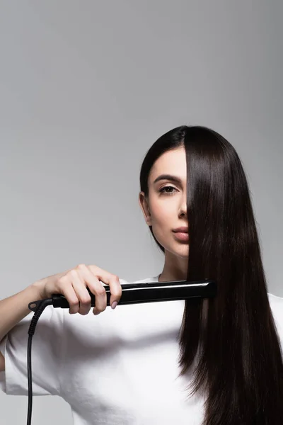 Joven morena mujer usando plancha de pelo aislado en gris - foto de stock