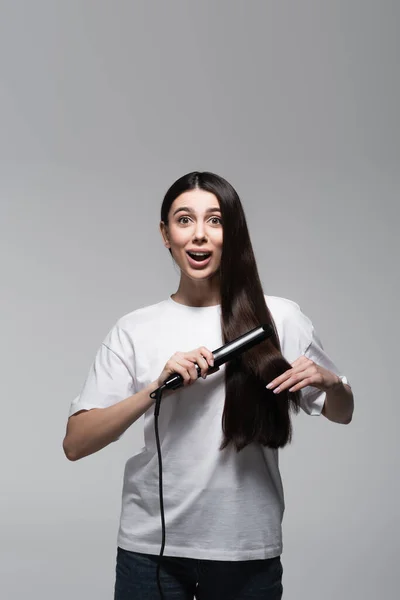 Jovem mulher surpreso usando alisador de cabelo isolado em cinza — Fotografia de Stock