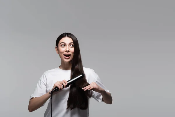Amazed woman using hair straightener isolated on grey — Stock Photo