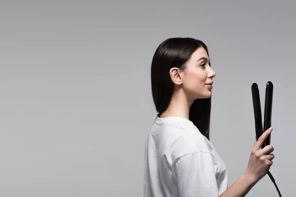 Side view of pleased woman holding hair straightener isolated on grey — Stock Photo