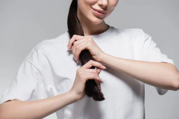 Vista recortada de mujer alegre en camiseta blanca tocando pelo castaño aislado en gris - foto de stock
