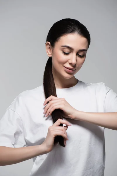 Fröhliche Frau im weißen T-Shirt, die braune Haare isoliert auf grau berührt — Stockfoto