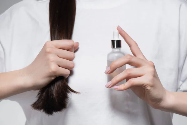Vista recortada de la joven mujer sosteniendo botella con aceite cerca de pelo brillante - foto de stock