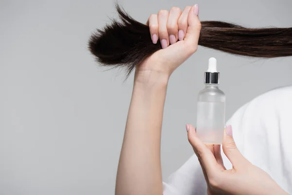Abgeschnittene Ansicht der jungen Frau mit Flasche mit Öl und glänzendem Haar isoliert auf grau — Stockfoto