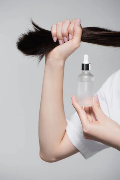 Vista parcial de una joven sosteniendo botella con aceite y pelo brillante aislado en gris - foto de stock